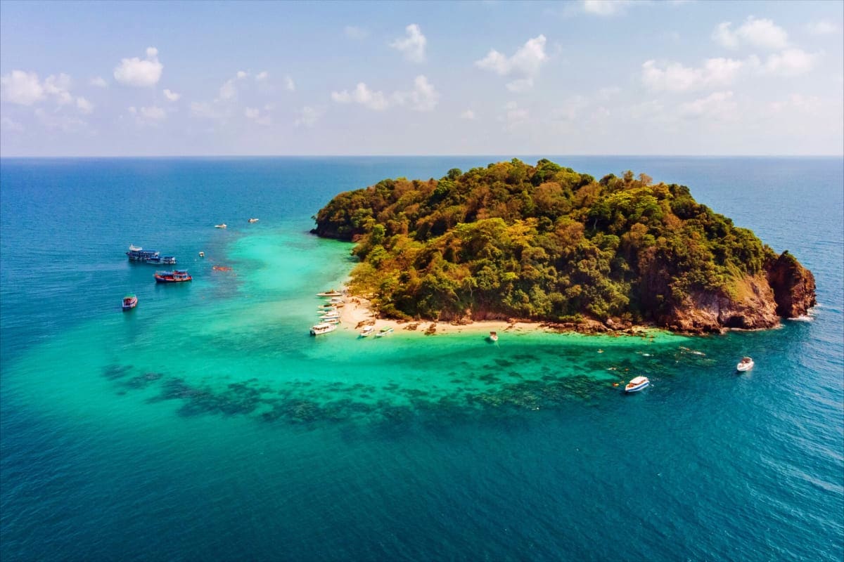 Tropical island with dense green vegetation, sandy beach, and turquoise waters surrounded by moored boats under blue sky with