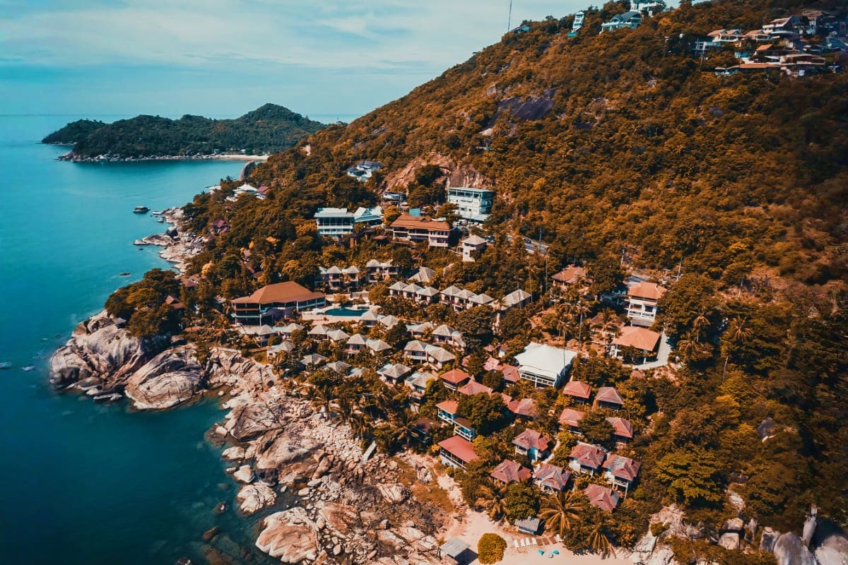 Aerial view of a coastal hillside village with red-roofed houses nestled among dense green vegetation overlooking turquoise M