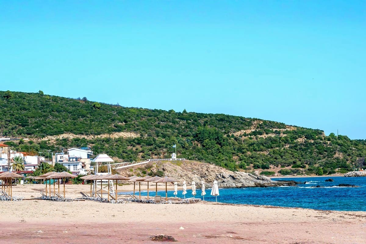 Mediterranean beach resort with sandy shore, blue water, wooden beach loungers, rocky hillside covered in green vegetation, a