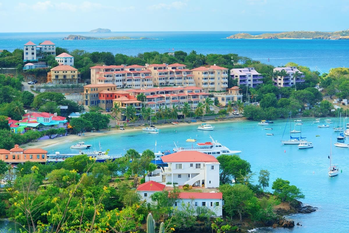 Coastal Greek island village with colorful waterfront buildings, sailboats in turquoise bay, and white houses with red-tiled 