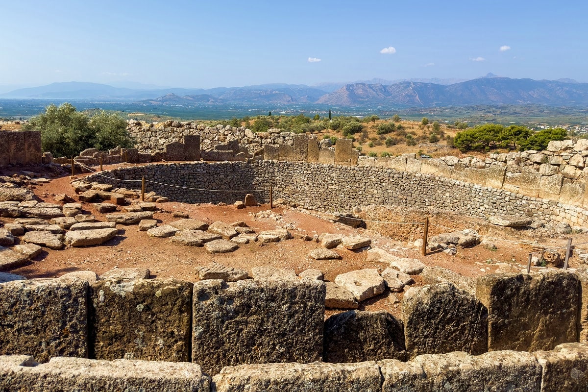 Ancient Greek ruins with stone walls and scattered columns overlooking a vast valley with mountains in the distance under cle