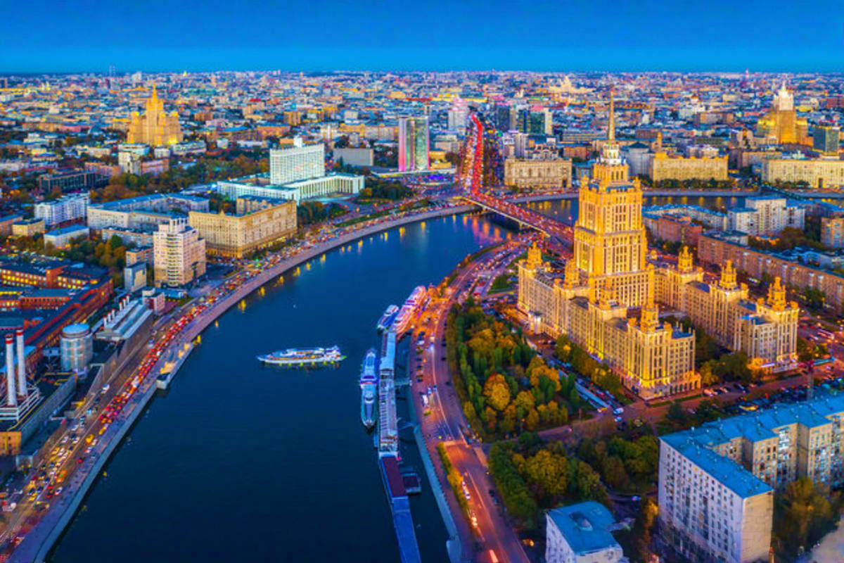 Aerial view of Moscow cityscape at dusk with illuminated Stalin-era skyscraper, Moscow River, and cruise ship docked at river