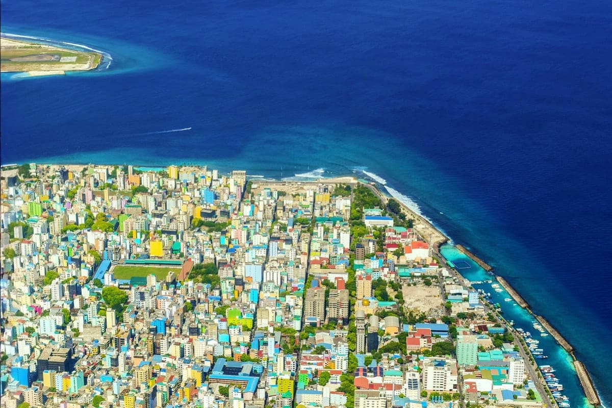 Aerial view of Male city in the Maldives with colorful buildings, a coral reef lagoon, and a distant island resort
