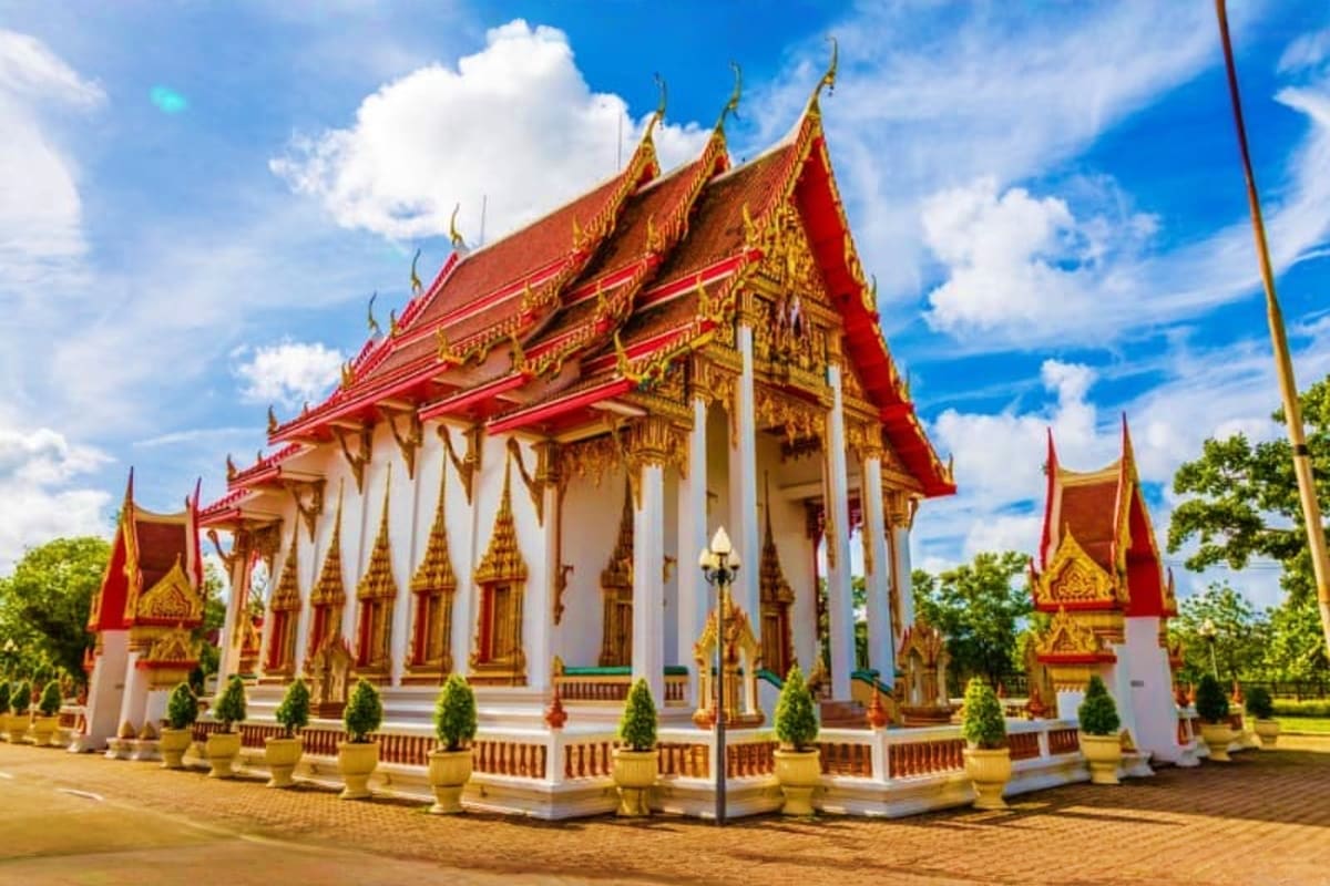 Traditional Thai Buddhist temple with red and gold roofs, white pillars, and ornamental spires against blue sky in Phuket