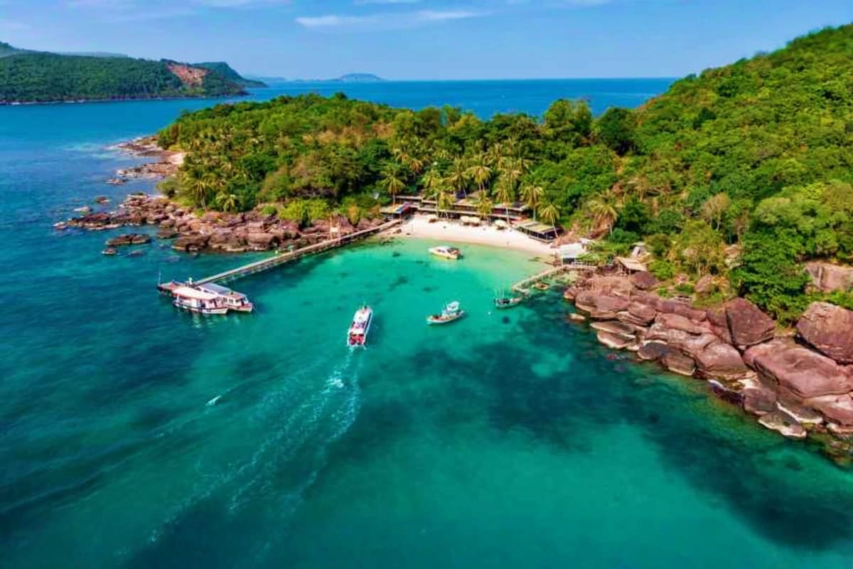 Aerial view of a tropical island cove with turquoise water, boats, white sand beach, wooden pier, and lush green vegetation s