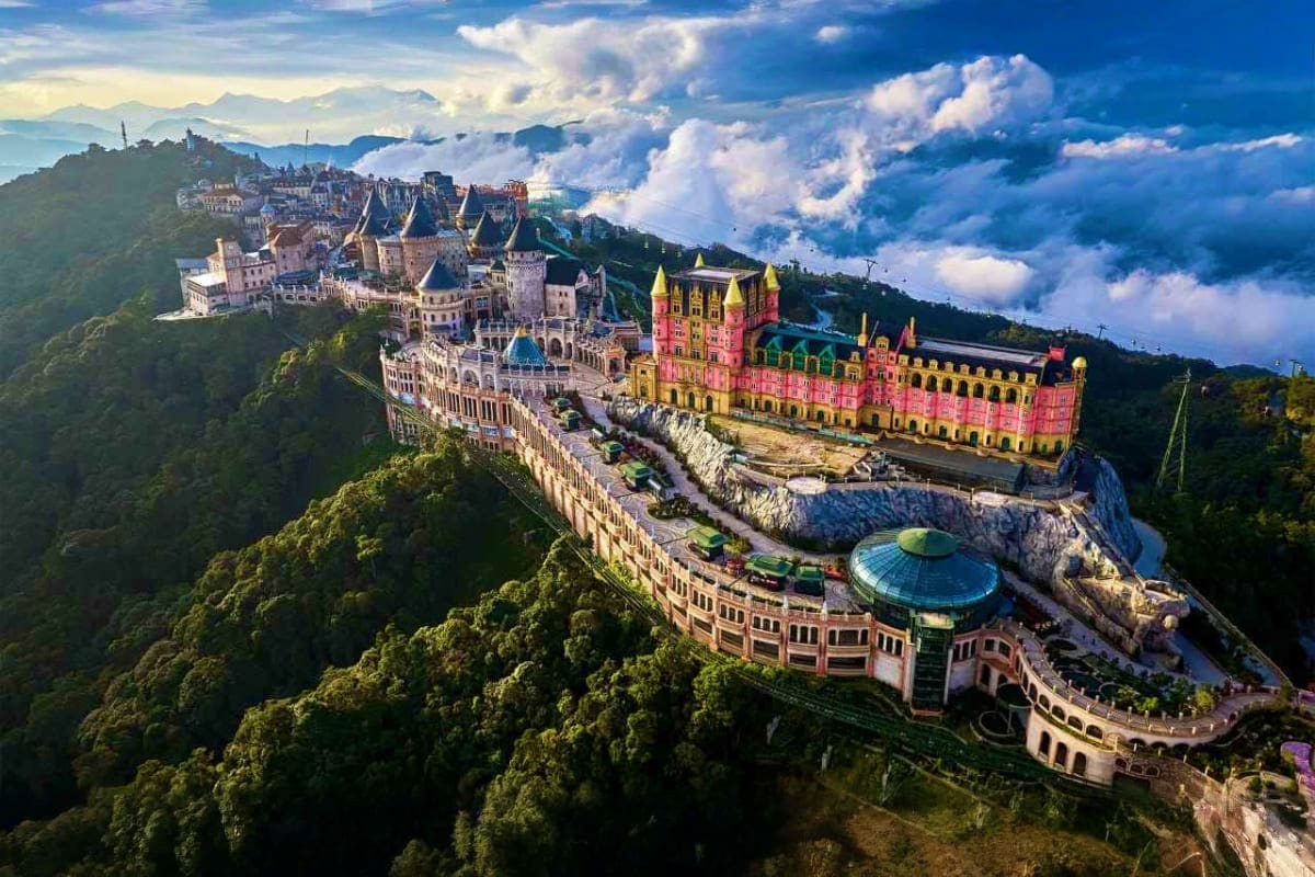Aerial view of a colorful castle resort with pink and yellow buildings nestled on a forested hilltop surrounded by clouds and