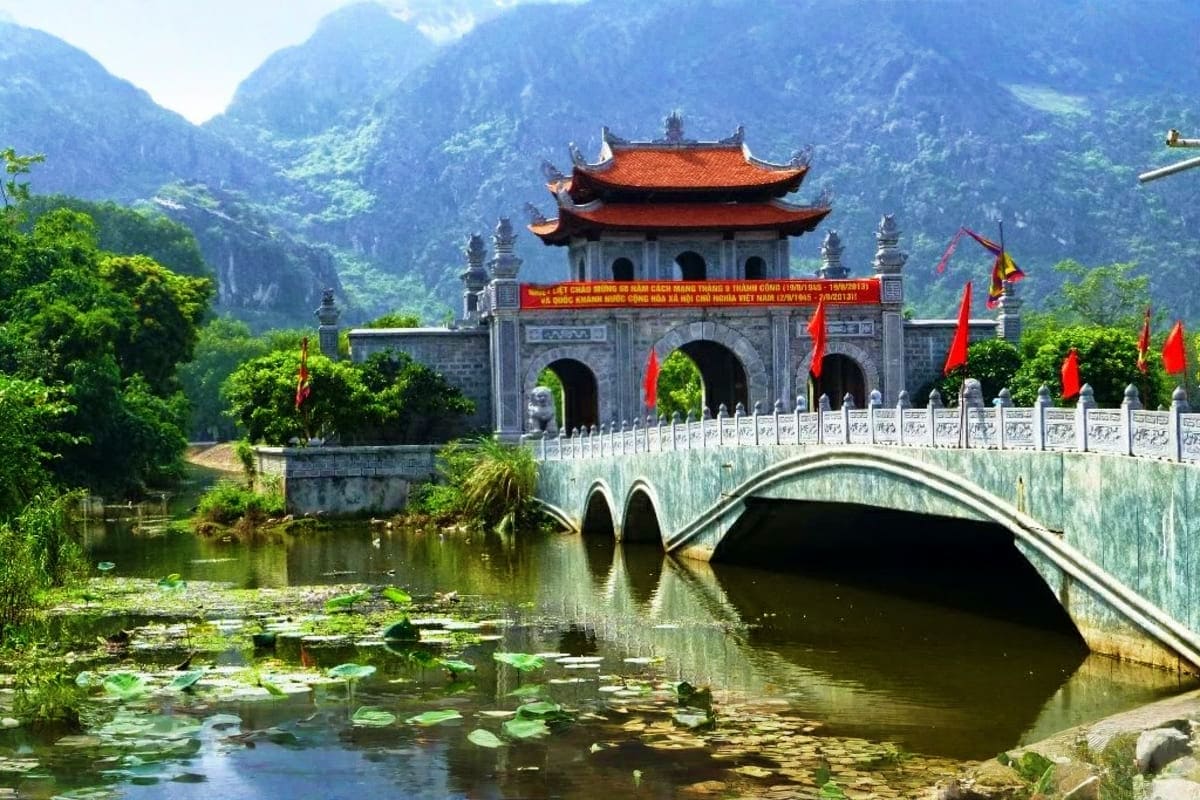 Ancient Vietnamese temple with red tiled roof and ornate stone gateway spanning across lily pond, surrounded by dramatic lime