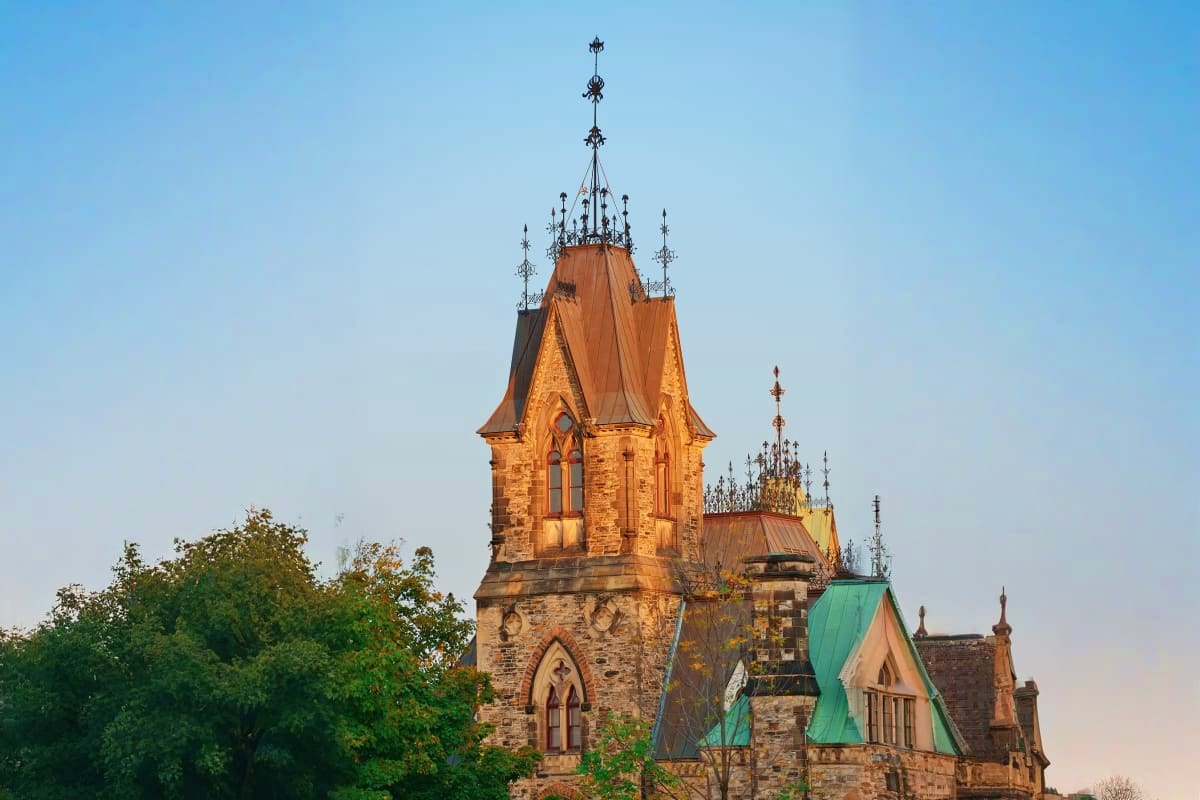 Gothic Revival stone tower with ornate spires and copper roofs against clear blue sky, surrounded by green trees