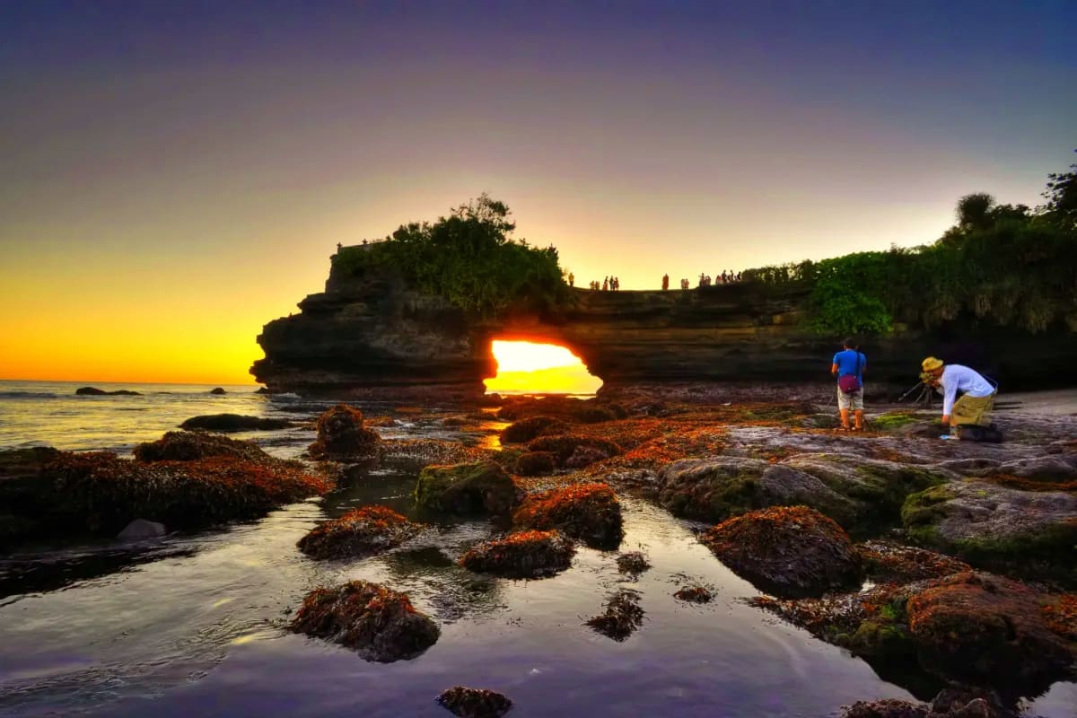 Sunset at Batu Bolong rock formation in Bali with tourists exploring tide pools and natural arch illuminated by golden light