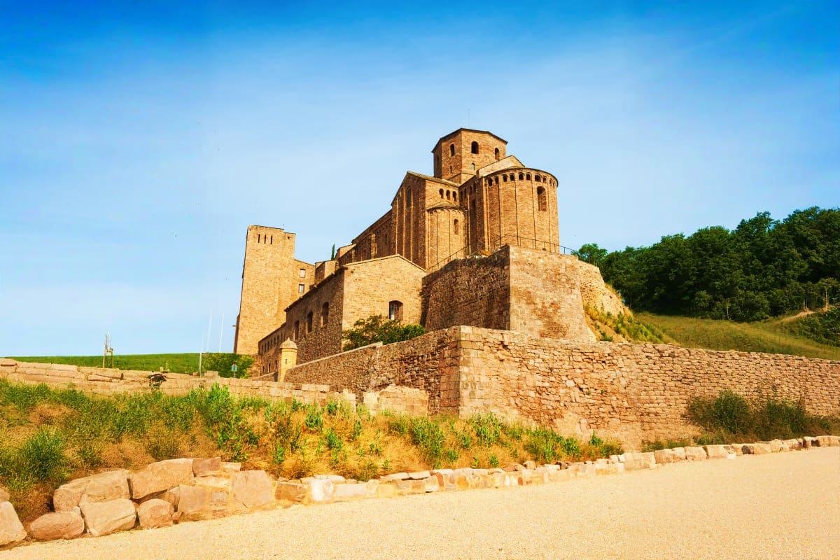 Medieval brick fortress with domed towers on hilltop surrounded by stone walls, green fields and forest under clear blue sky
