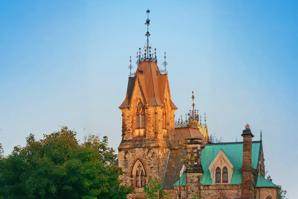 Gothic Revival stone church tower with ornate spires and green copper roof against clear blue sky, surrounded by green trees