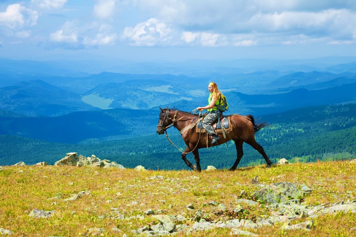 Woman riding brown horse on mountain meadow overlooking forested valleys and lakes in Georgia