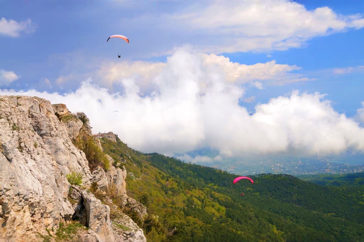 Paragliders soaring above forested mountains and rocky cliffs in Georgia with white clouds and blue sky