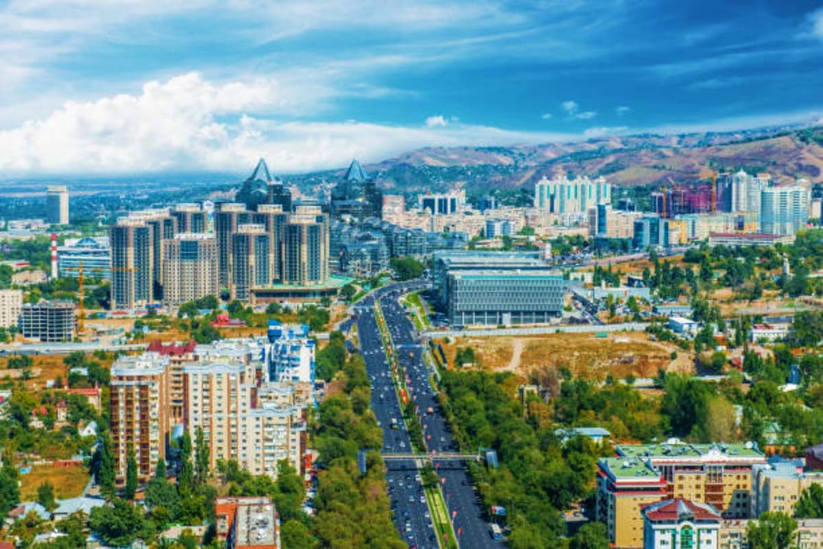 Aerial view of Almaty cityscape with modern high-rise buildings, mountains in background, and wide avenue with traffic