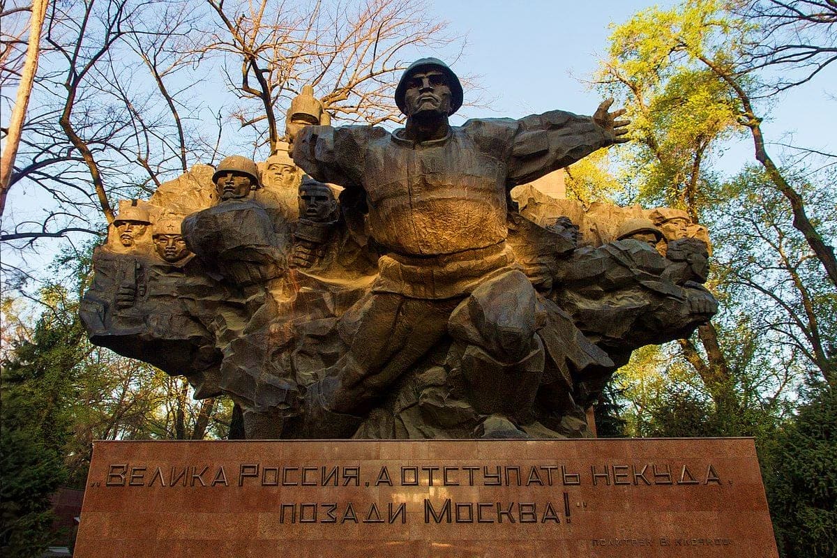 Bronze monument of Soviet soldiers with central figure with raised arms, surrounded by bare trees and green foliage, with Cyr