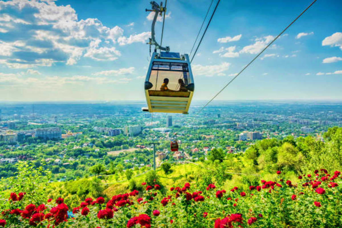 Aerial cable car ride over mountainous landscape with red flowers, green forests, and city sprawl below in Kazakhstan