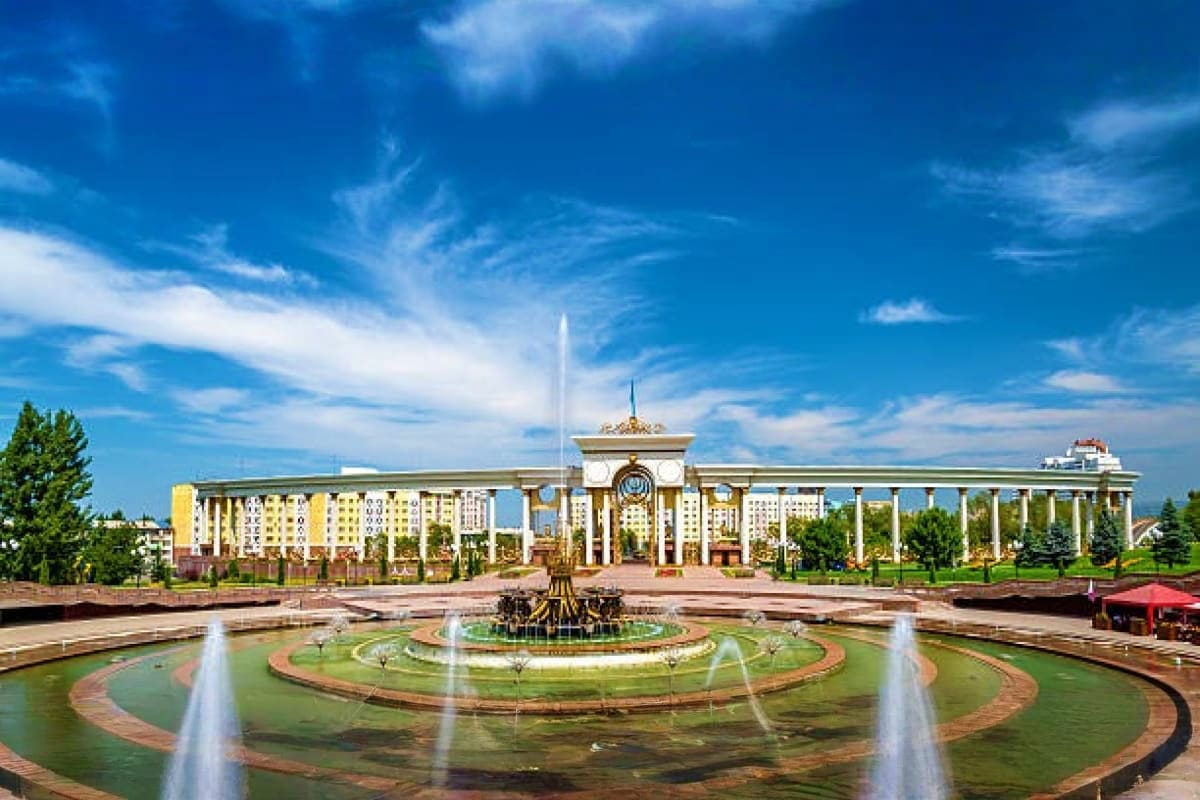 Central plaza with ornate fountain and water features in front of grand government building with columns under blue sky, Asta