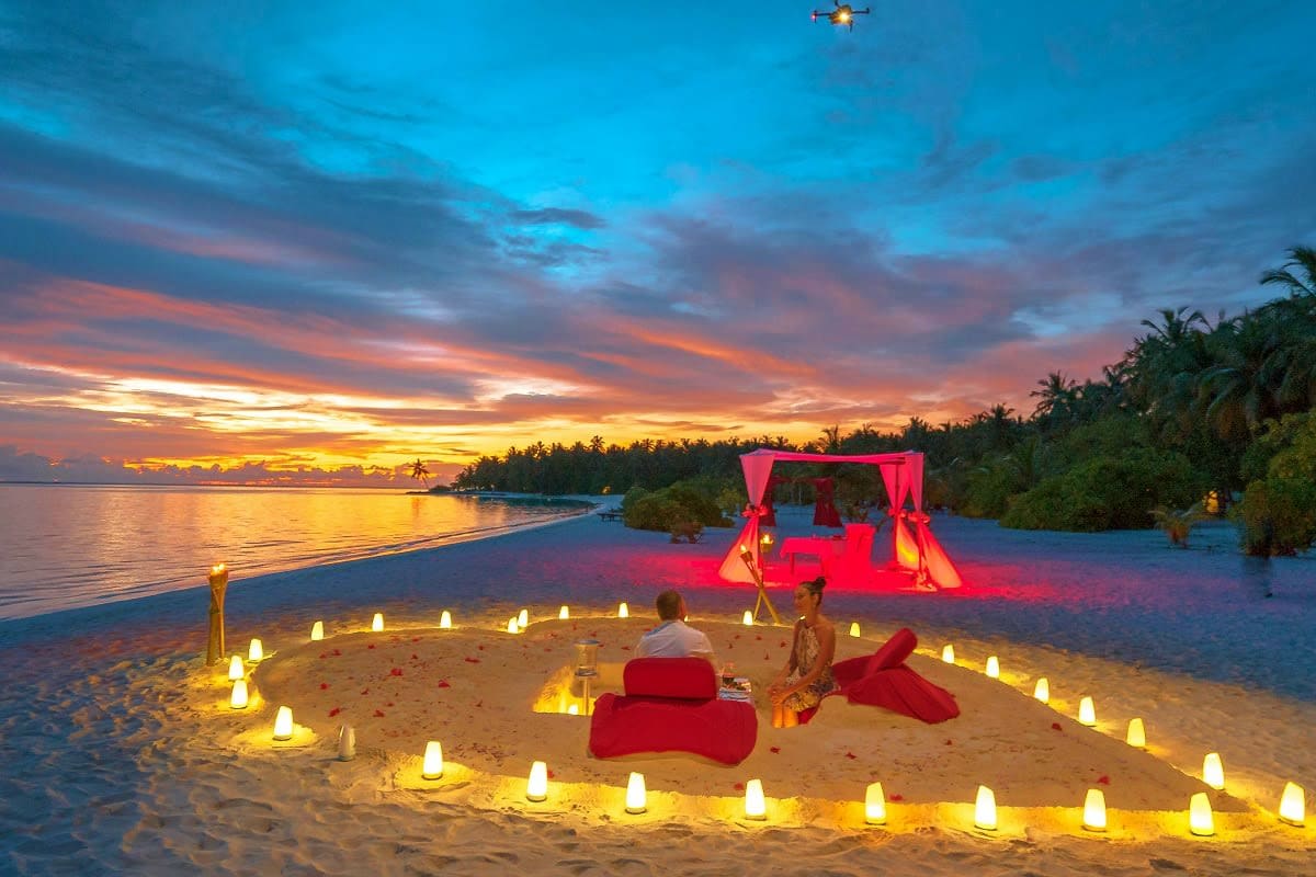 Romantic beach setup at sunset with couple sitting on sand surrounded by lit candles, red fabric decorations, and illuminated