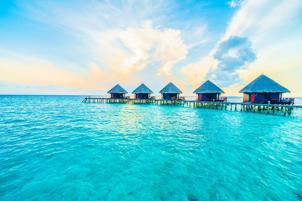 Row of overwater bungalows with thatched roofs in turquoise tropical water under blue sky with white clouds, Maldives