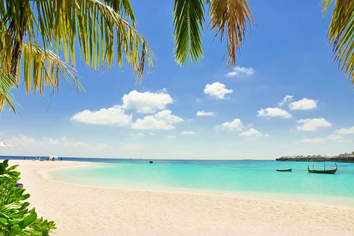 Tropical beach with turquoise water, white sand, palm fronds, moored boats, and blue sky with white clouds