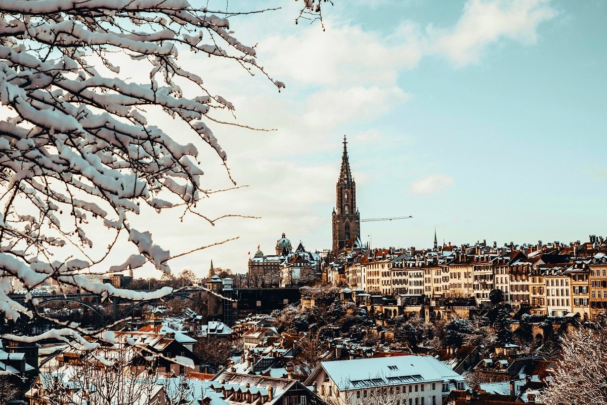 Snow-covered tree branches frame a winter view of Bern's historic cityscape with the iconic Cathedral spire rising above trad