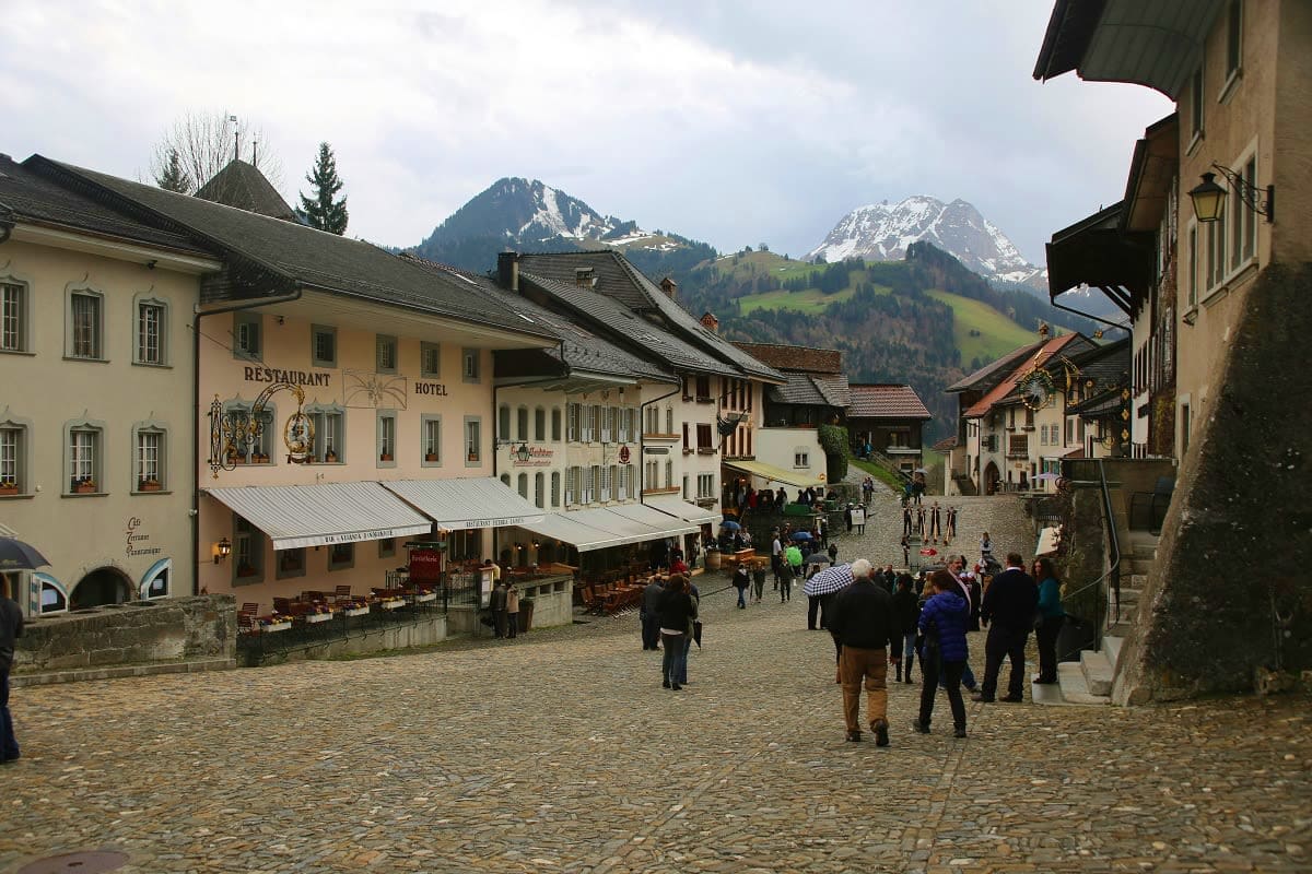 Medieval Swiss alpine village square with cobblestone plaza, traditional chalets, restaurant and hotel buildings, snow-capped