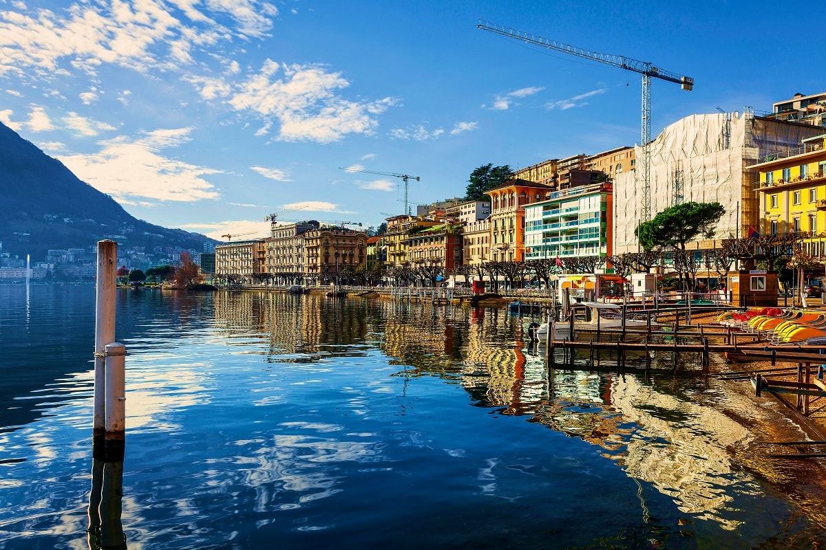 Lakeside waterfront promenade in Geneva with colorful buildings, construction cranes, and snow-capped mountains reflected in 