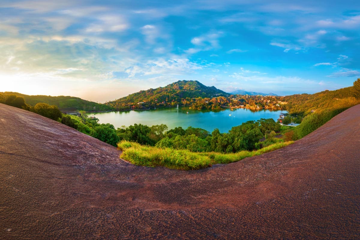 Scenic lake view surrounded by forested hills with blue water, mountain peak, residential buildings, and clear sky at sunrise