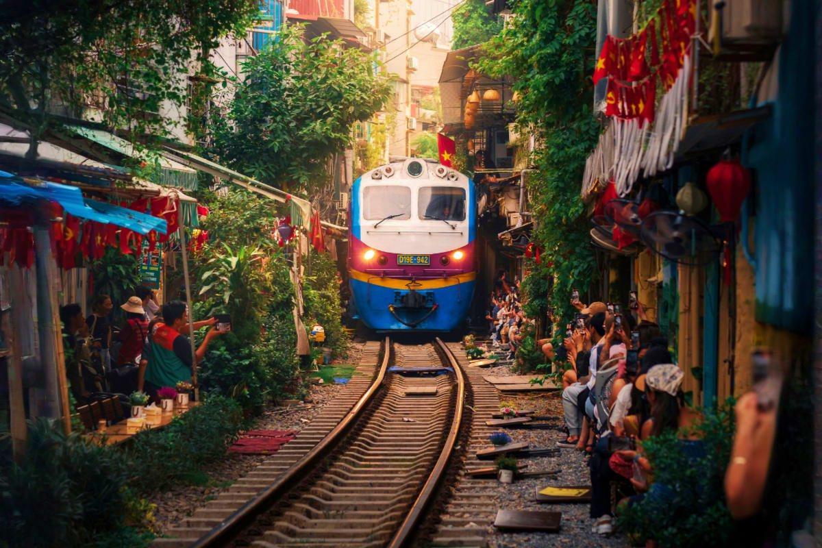Blue and white train moving through narrow Hanoi street lined with buildings, greenery, and local residents sitting along the
