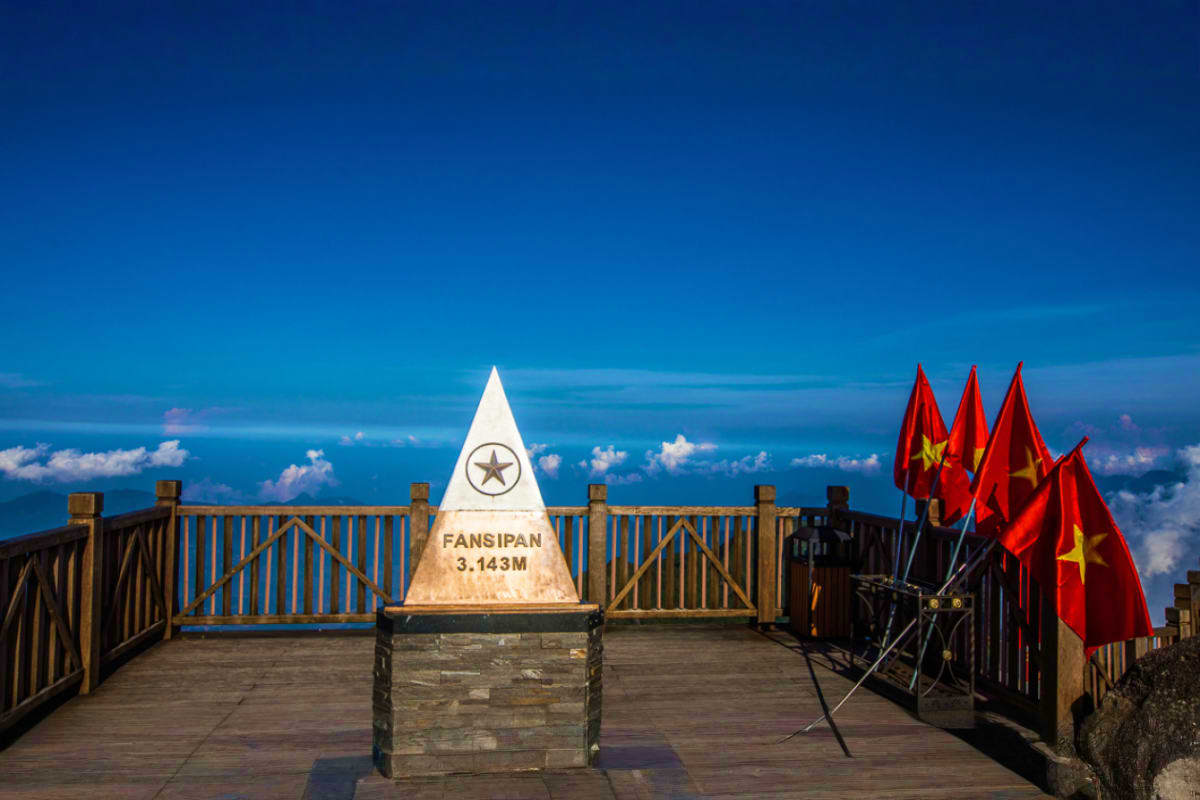Fansipan peak marker at 3,143 meters with Vietnamese flags on wooden observation deck overlooking clouds and blue sky