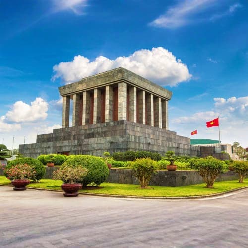 Ho Chi Minh Mausoleum in Hanoi with Vietnamese flags, surrounded by manicured gardens and blue sky with white clouds