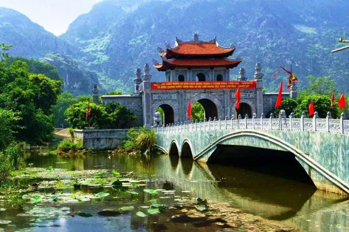 Ancient stone temple with red curved roof and ornate gates beside arched bridge over tranquil river in mountainous Vietnamese