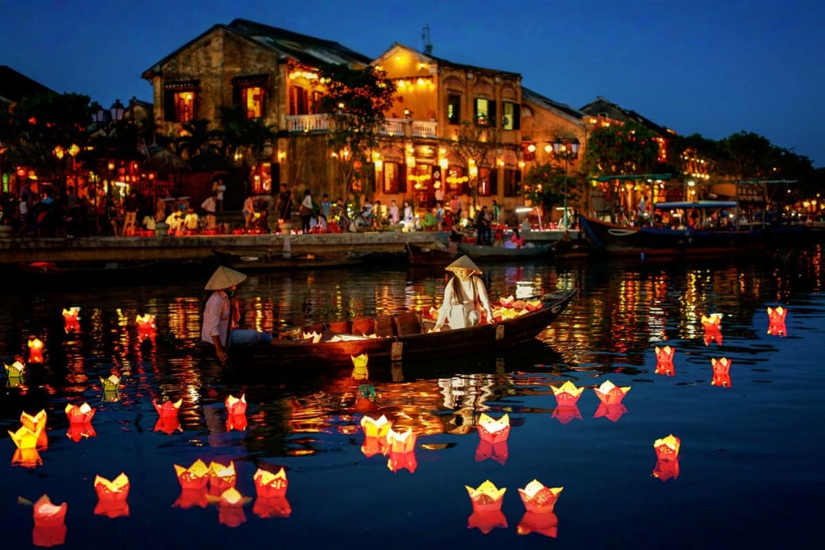 Hoi An ancient town at twilight with illuminated yellow buildings, wooden boats, and colorful floating lanterns on the river