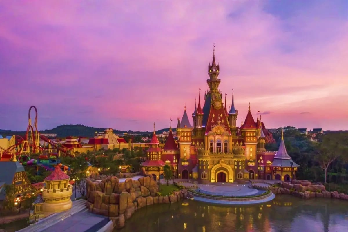 Illuminated fantasy castle at twilight with purple sky, surrounded by theme park attractions and water feature at dusk