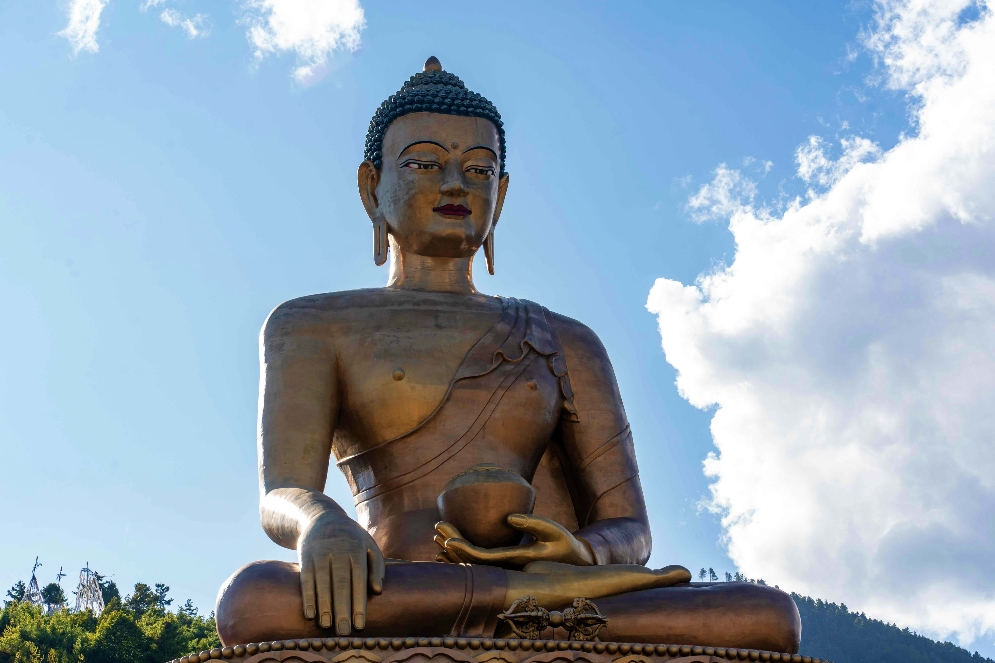 BHUTAN Seated bronze Buddha statue holding a scepter against blue sky with white clouds and forested mountains in background, Bhutan