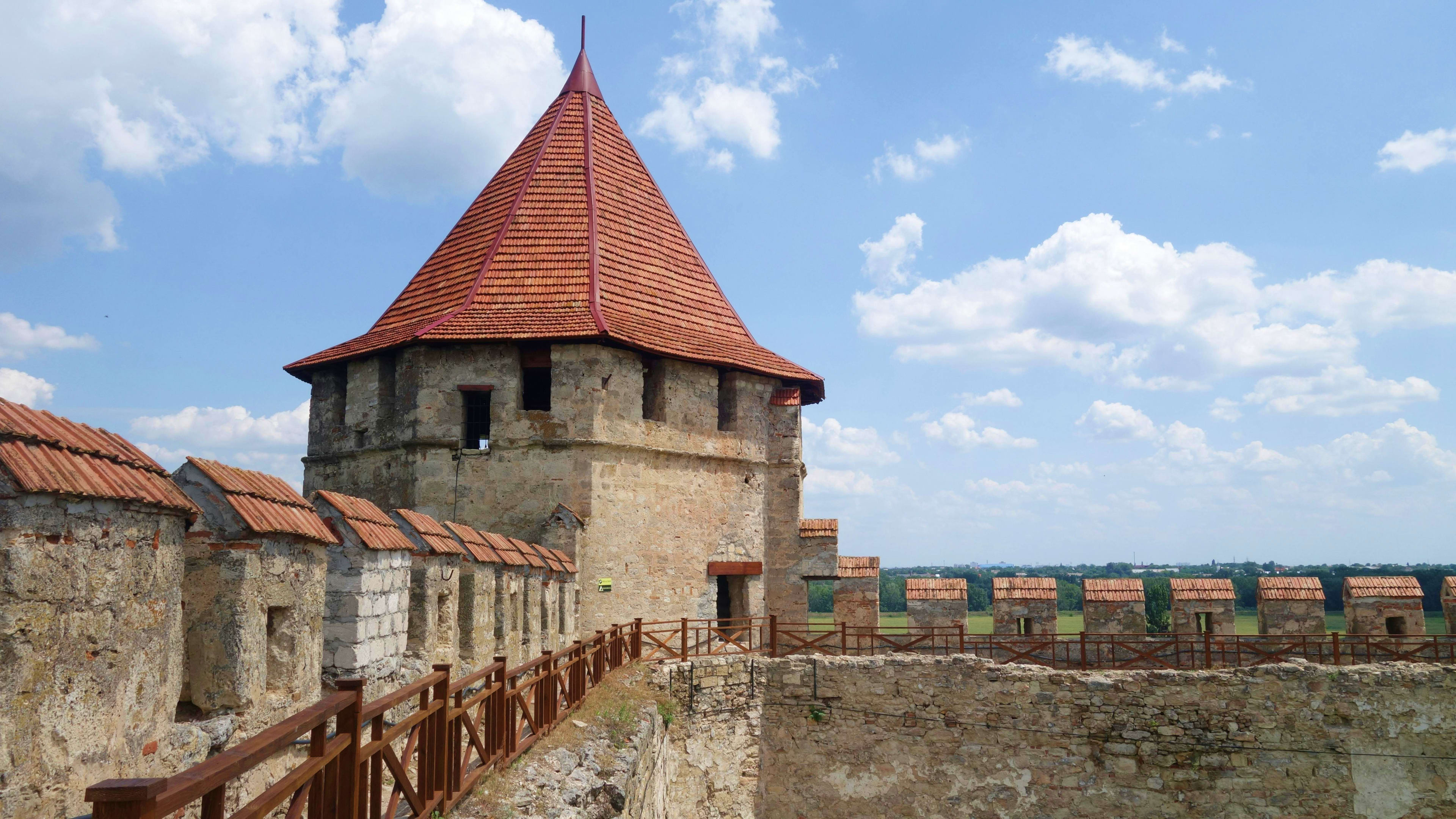 Moldova Medieval stone fortress with conical red-tiled tower, wooden walkway, and green countryside landscape under blue sky in Moldo