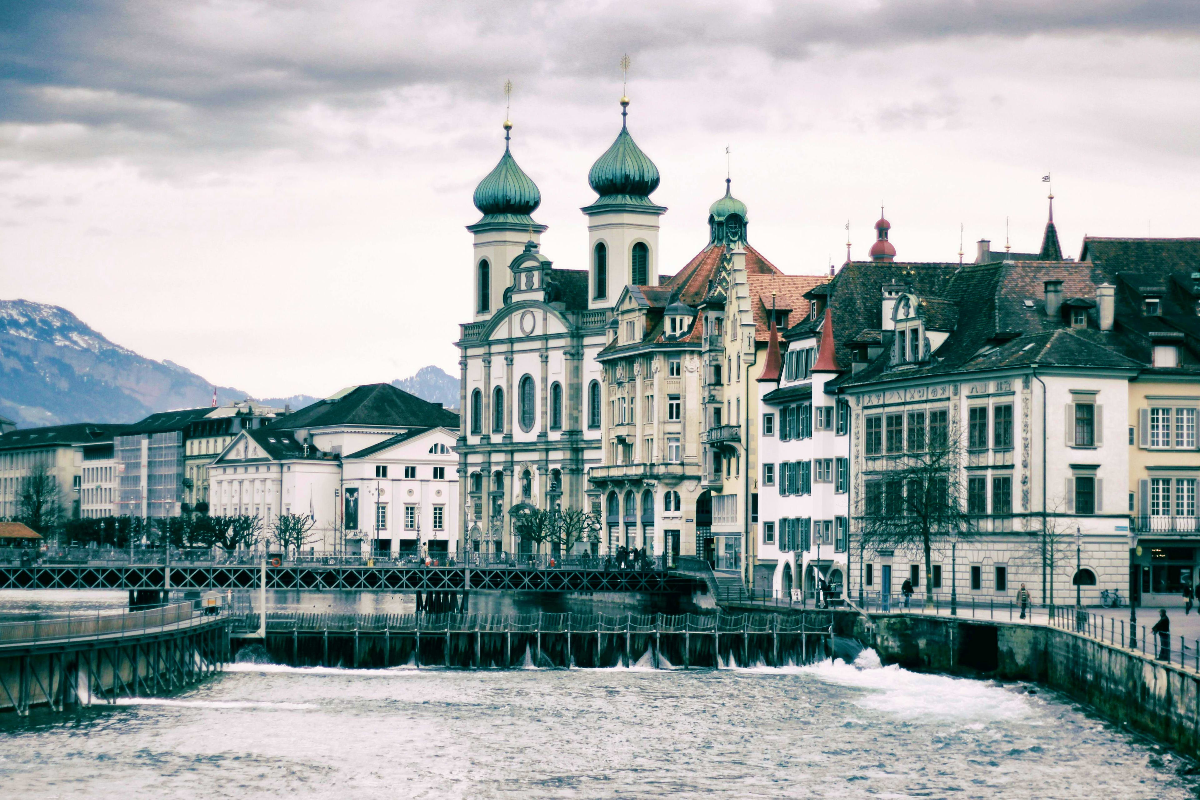Switzerland Historic Lucerne cityscape with white church featuring green onion domes reflected in the Reuss River, mountains in backgroun
