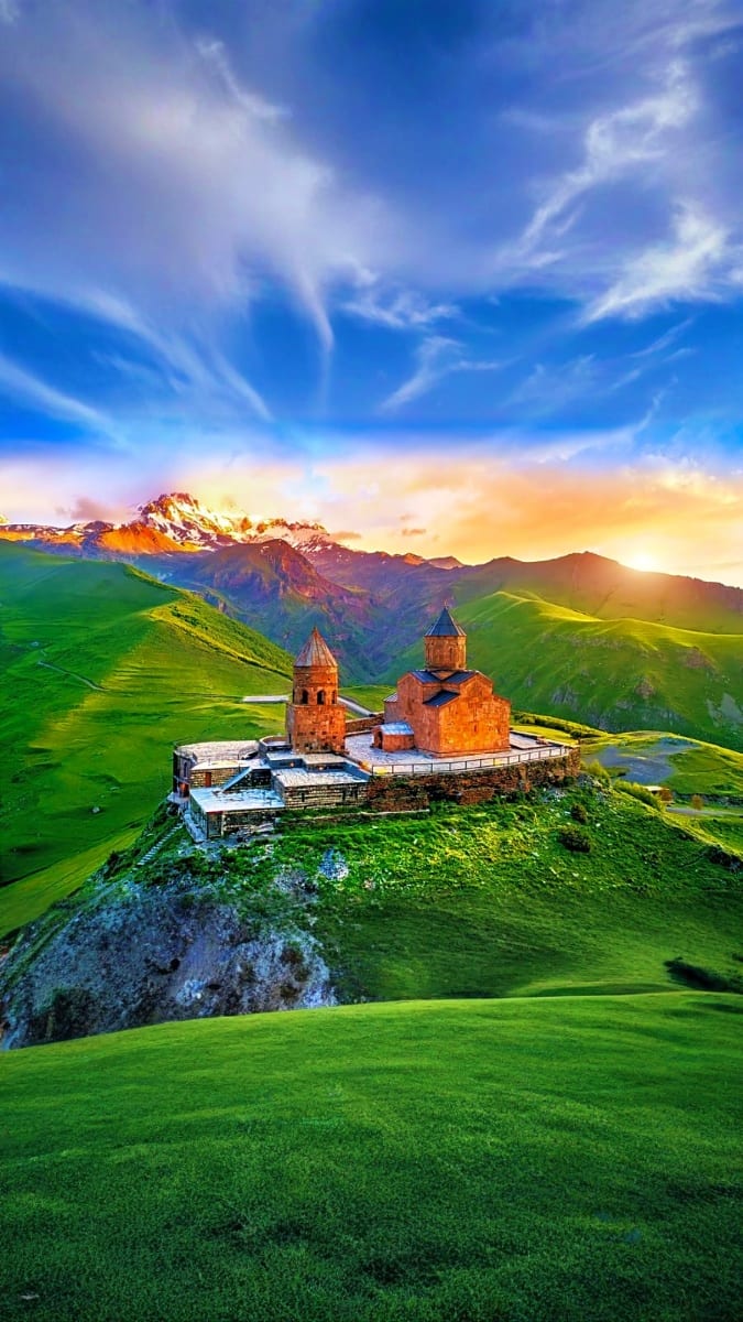 Svetitskhovleba church complex at sunset with snow-capped mountains and green rolling hills in Georgia