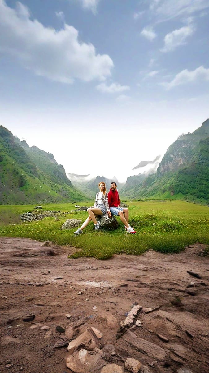Couple sitting on a rock in a lush alpine valley surrounded by green mountains and mist in the Caucasus region of Georgia