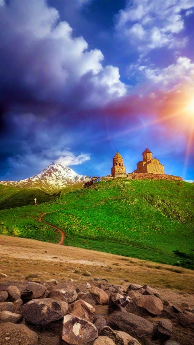 Ancient Armenian church on green hillside with snow-capped Mount Ararat in background under dramatic cloudy sky