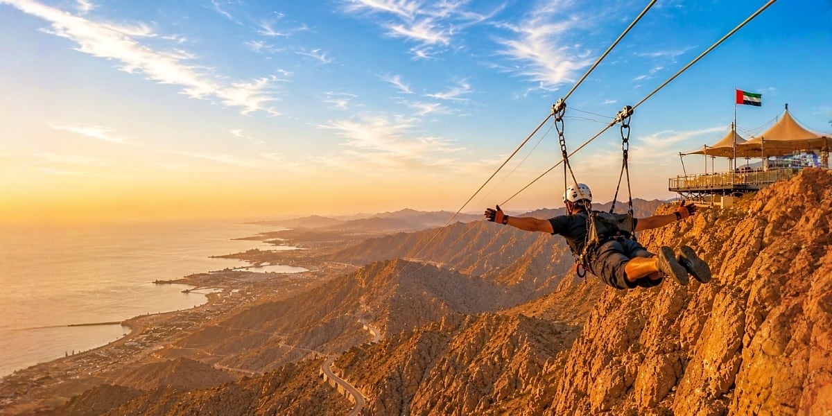 Person ziplining off Jebel Jais mountain with coastal valley and UAE flag visible at sunset