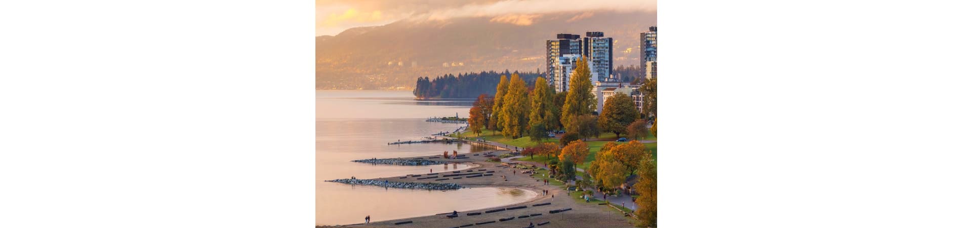 Vancouver waterfront at sunset with high-rise buildings, beach, autumn trees, and North Shore mountains