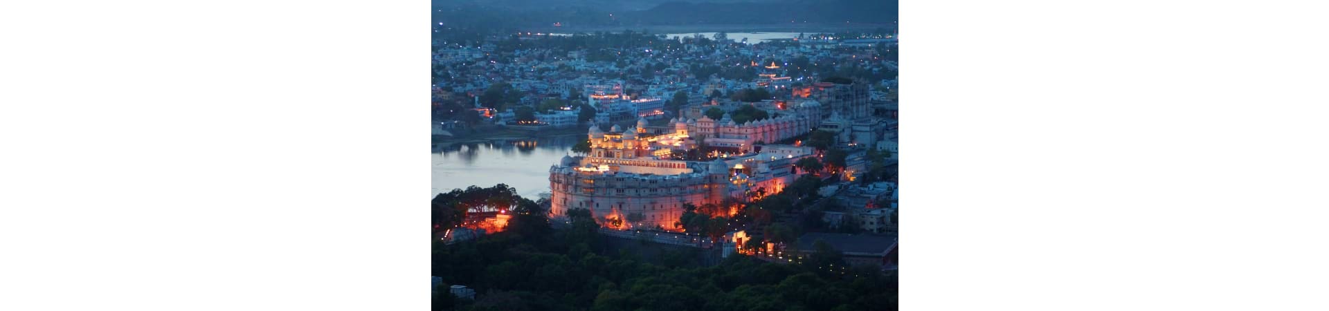 Udaipur cityscape at dusk with illuminated palace on hilltop overlooking lake and surrounding buildings