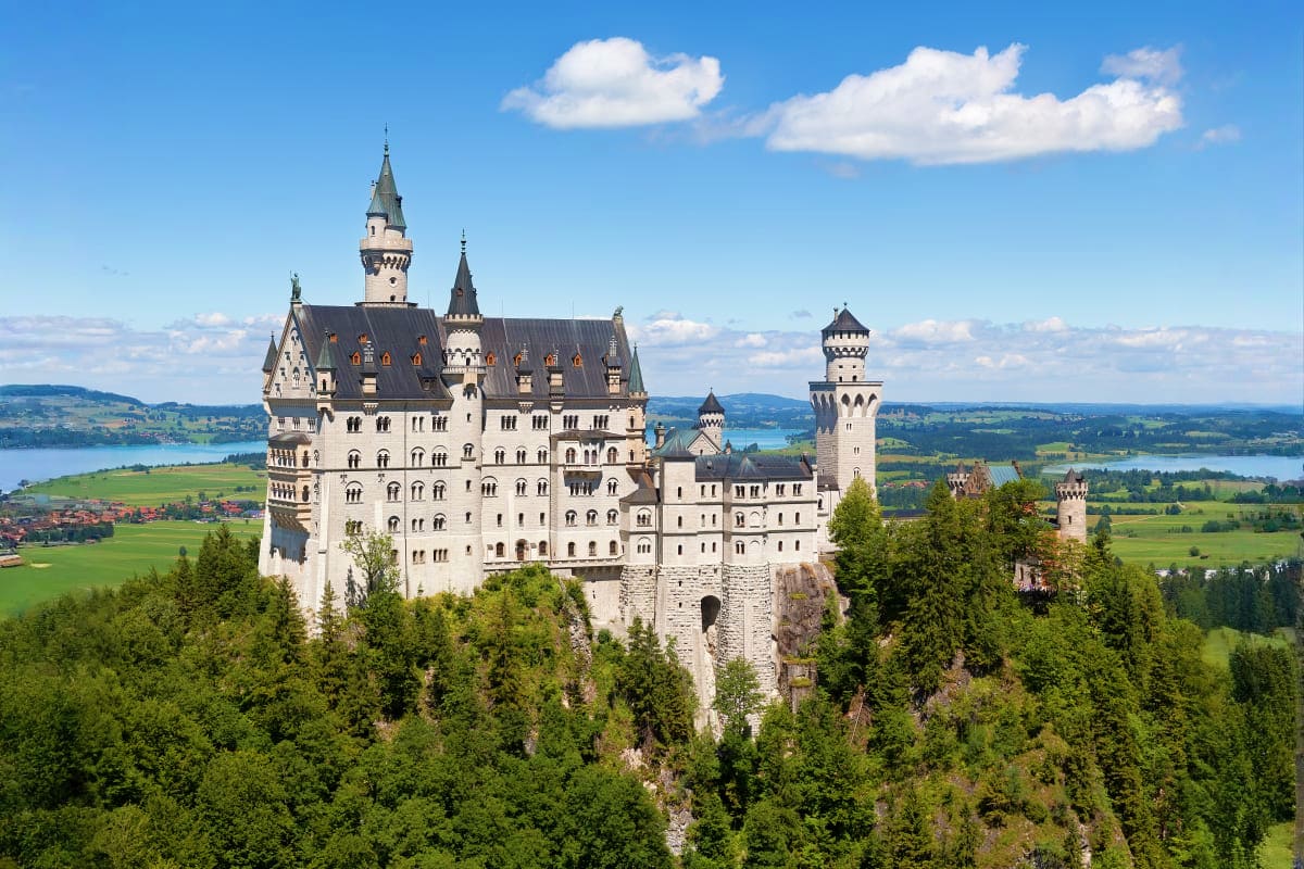 Neuschwanstein Castle perched on a rocky hilltop surrounded by forested mountains and Bavarian countryside with lakes in the 