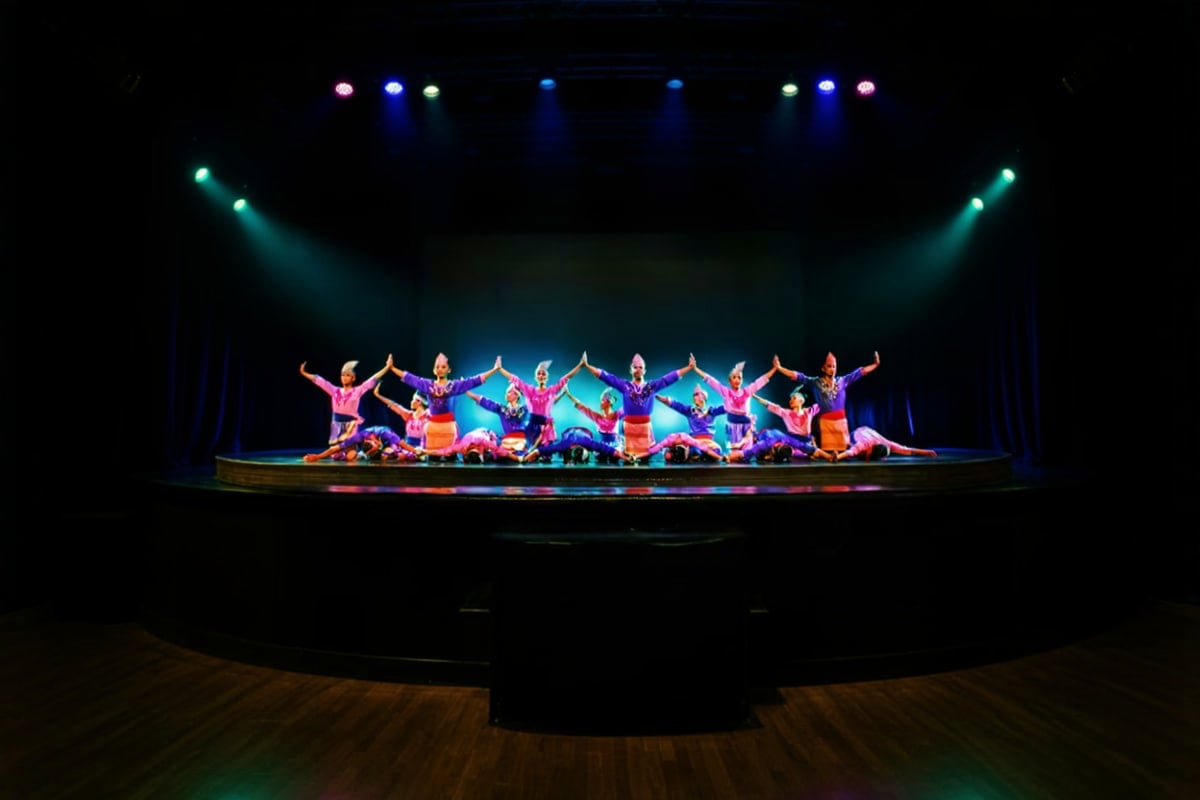 Large ensemble of performers in colorful pink and blue costumes dancing on stage with synchronized poses and raised arms unde