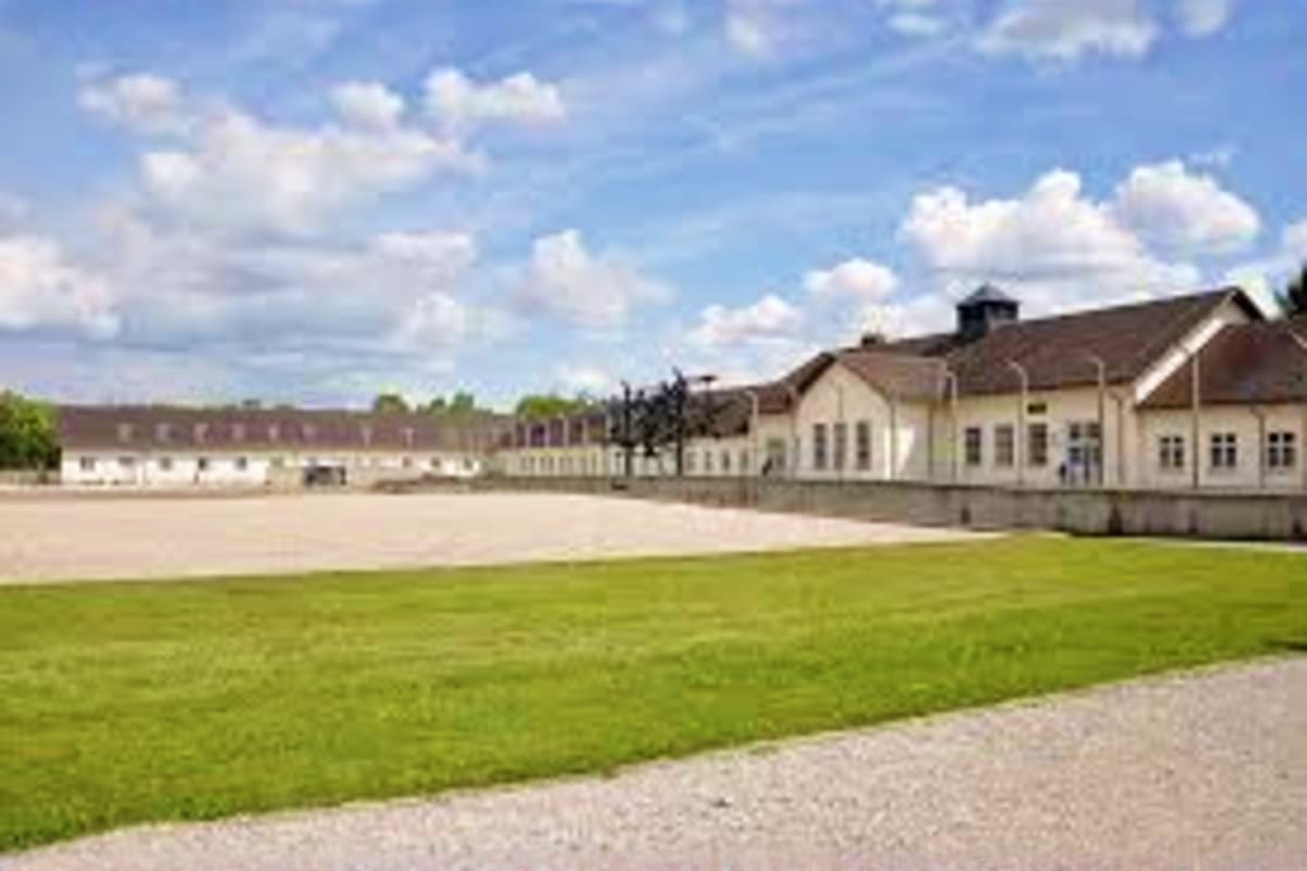 Dachau concentration camp memorial site with cream-colored barracks buildings, green lawn, gravel path, and cloudy sky