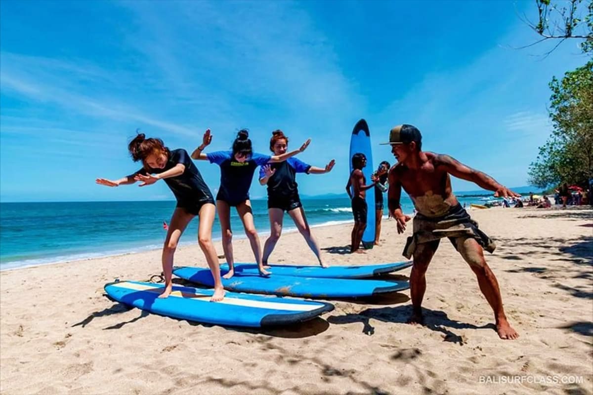 Surf instructor teaching children surfing lesson on blue boards at Kuta Beach with ocean and sandy shore in background
