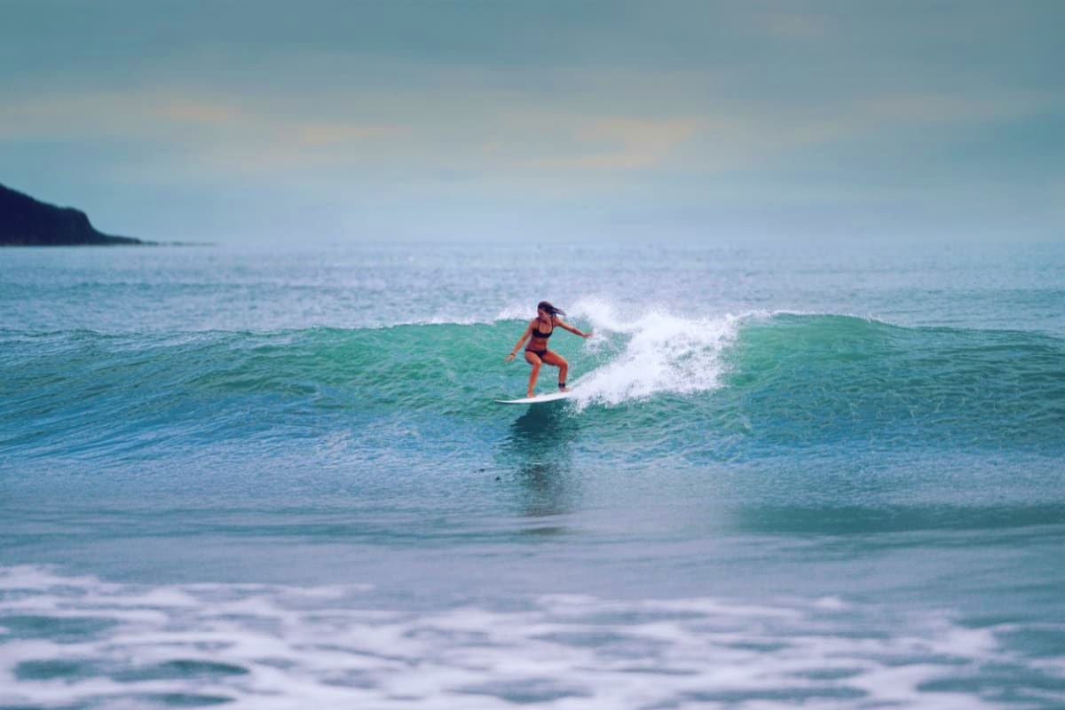 Woman surfing on a green wave at Kuta Beach with cliffs visible in the background under a cloudy sky