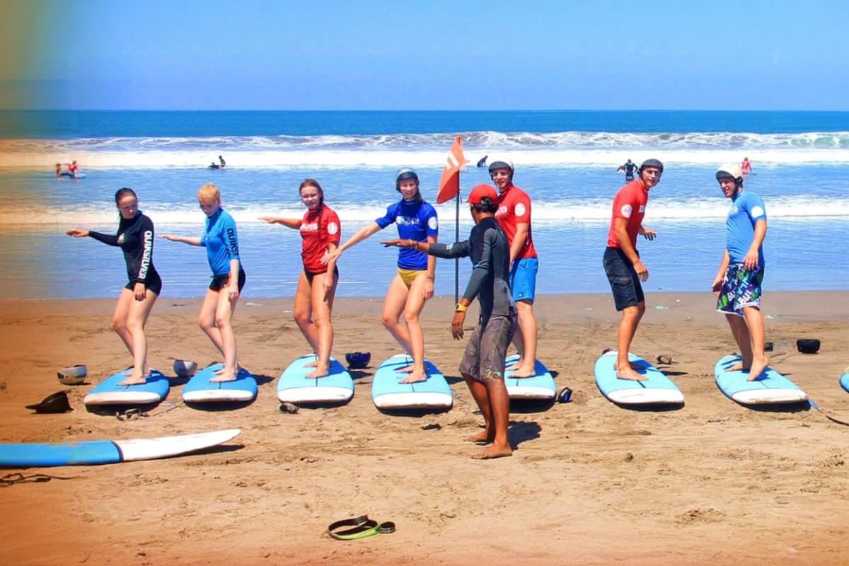 Group of surf students standing on blue surfboards on Kuta Beach sand with instructor demonstrating technique, ocean and wave