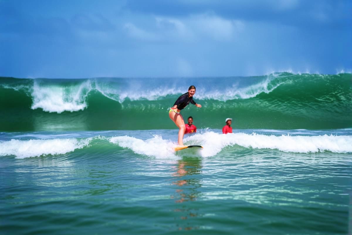 Female surfer riding a wave on yellow surfboard at Kuta Beach with instructors in red shirts watching in the background